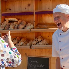 regionaler Bäcker Heller lacht mit einer Besucherin auf dem Regionalfest, im Hintergrund Baguettes in einer Auslage