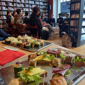 Snacks und belegte Brote auf zwei Tablets stehen auf einem Tisch, im Hintergrund sitzt eine Gruppe von Menschen