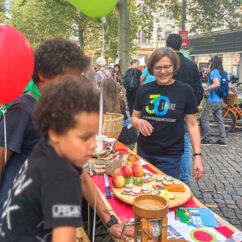 Kinder quetschen Bio-Getreide an einem Tisch, dahinter Mitarbeiterin der VG-Dresden