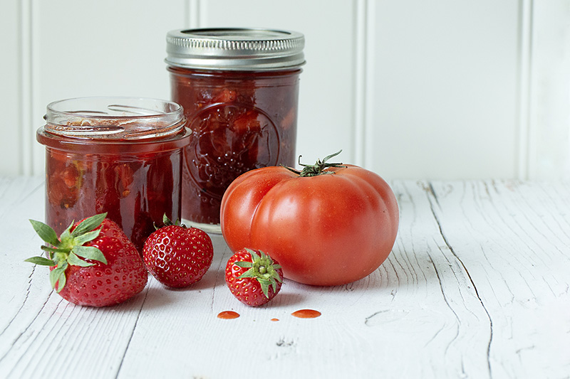 Zwei Gläser Erdbeer-Tomaten-Marmelade auf weißem Holzuntergrund davor drei Erdbeeren und eine Tomate