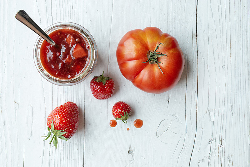 Ein Glas Tomaten-Marmelade mit Erdbeeren, daneben drei Erdbeeren und eine Tomate auf weißem Holzuntergrund