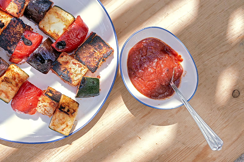 Drei Grill-Spieße mit mediterranem Gemüse und mariniertem Tofu, daneben Tomaten-Dip in einer Schale
