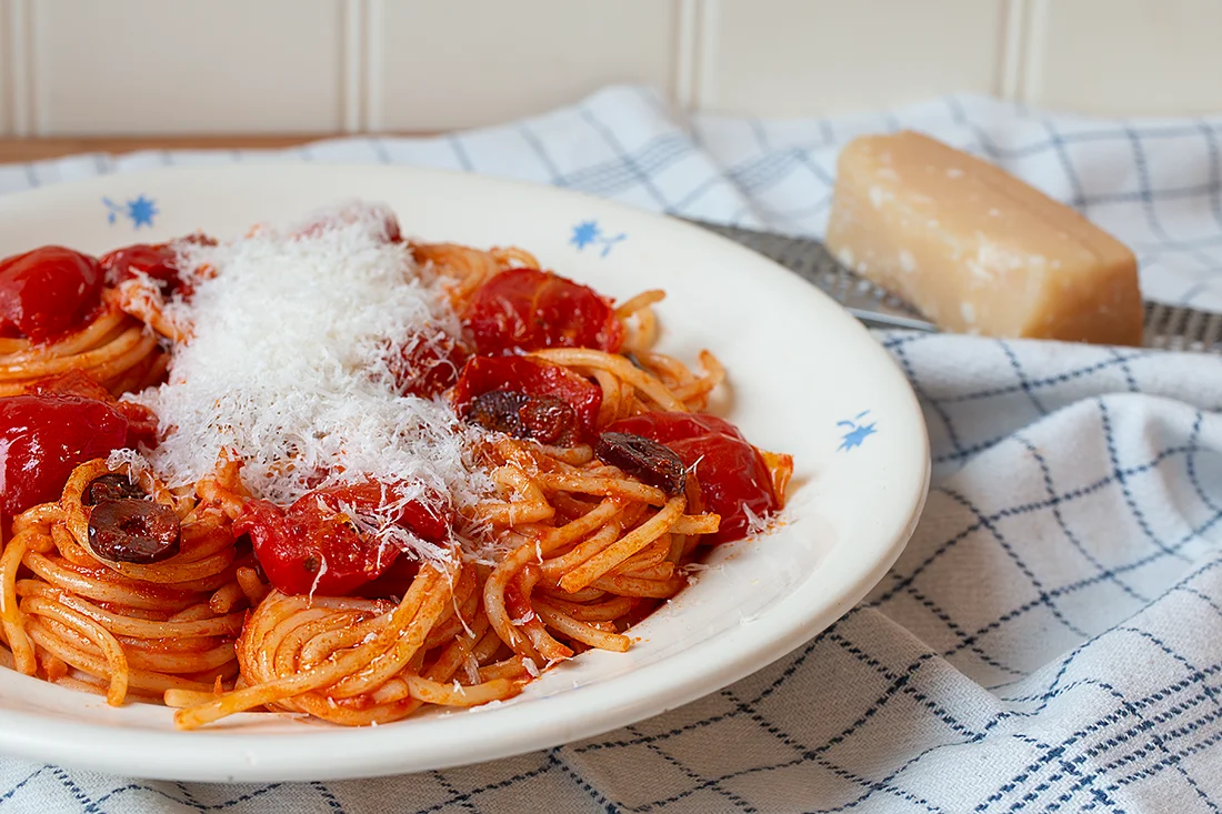 Spaghetti mit Tomaten, Oliven, Kapern und Käse auf einem Teller, daneben Käse und eine Käsereibe