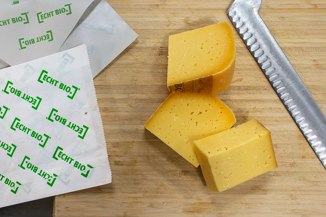 Käsepapier und Käsetüte liegen neben geschnittenem Käse und Messer auf einem Brett