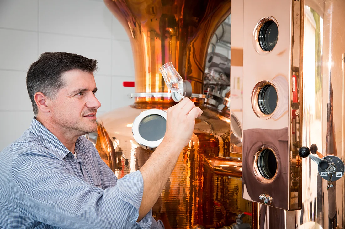 Mann steht vor einer Destillationsanlage und betrachtet eine Flüssigkeit im Glas prüfend gegen das Licht. Bild: Lautergold Paul Schubert GmbH